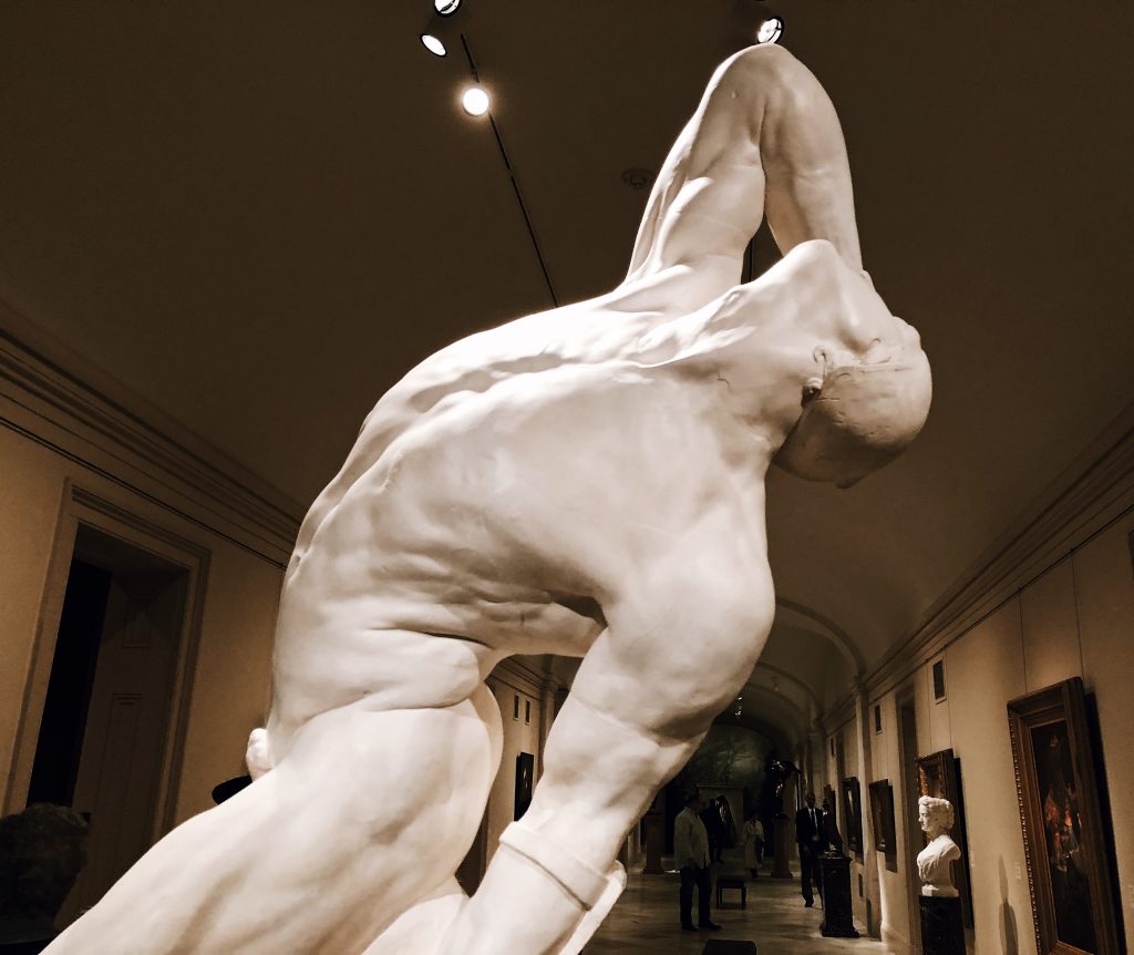 A white marble statue depicting a muscular figure arched backward with arms extended and head tilted back. The sculpture is displayed in a grand hallway with artworks on the walls and a softly lit ceiling.