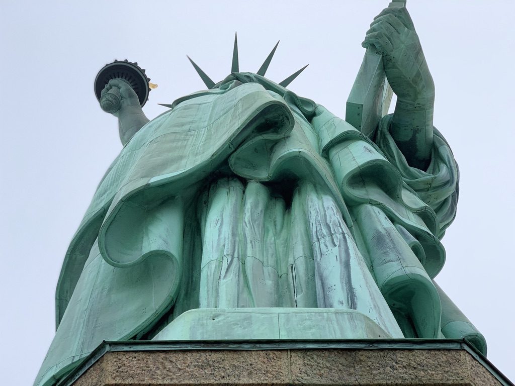Low-angle view of the Statue of Liberty, highlighting the folds of its robes and iconic torch. The statue is set against a cloudy sky, emphasizing its grand stature and architectural details.