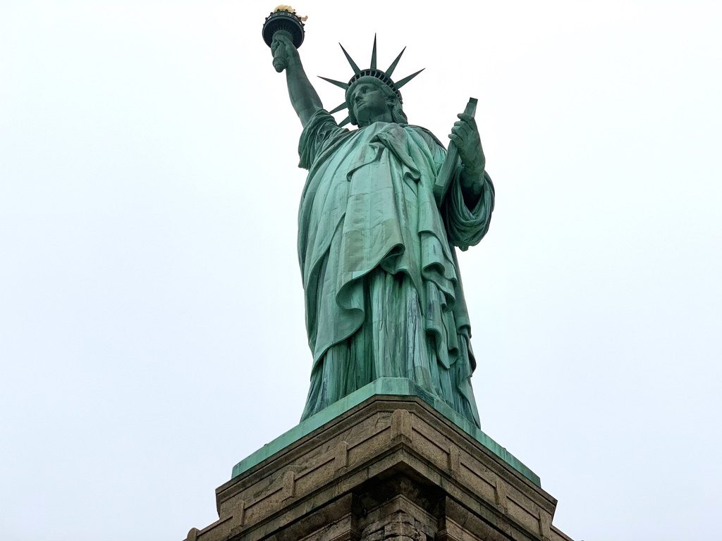 View of the Statue of Liberty from below, showcasing its iconic green patina, torch, and crown against a cloudy sky. The statue stands on its stone pedestal.