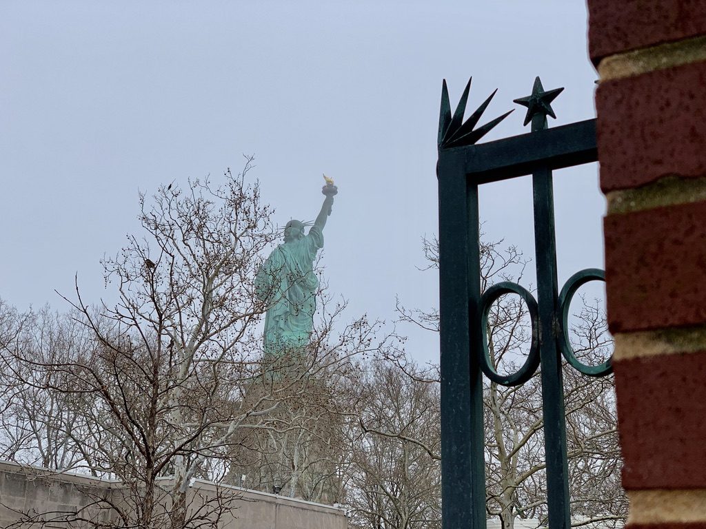 The Statue of Liberty partially obscured by bare winter trees and a decorative fence in the foreground on a foggy day.