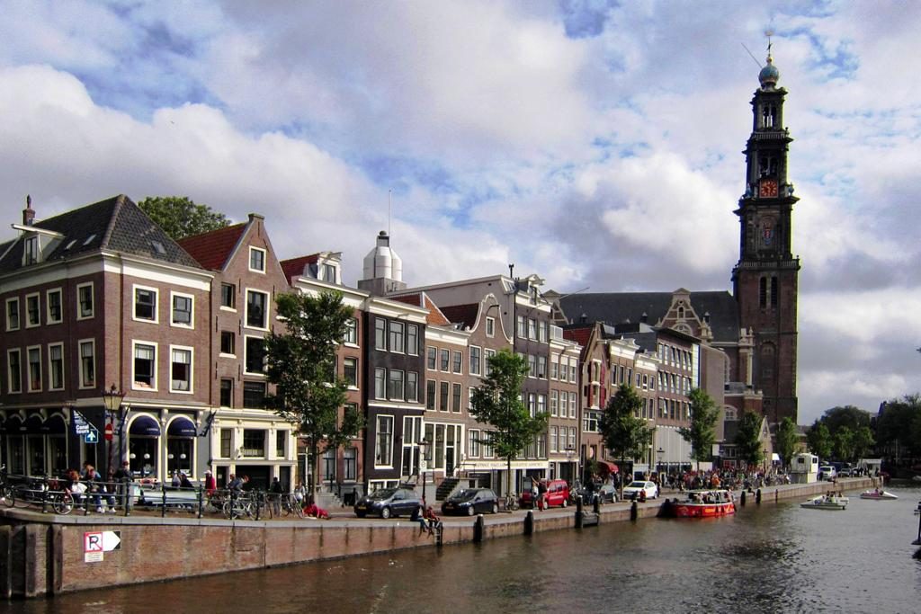 A scenic view of a canal in Amsterdam, lined with traditional Dutch buildings on the left. Bicycles are parked along the canal, with a cloudy sky above. A prominent tower stands in the background.