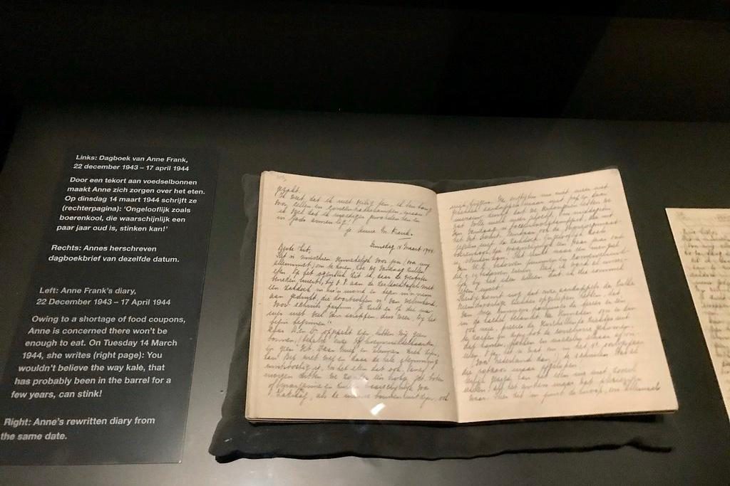 An open diary with handwritten text lies on display in a glass case. A description in Dutch is shown on the left, and an English translation is provided underneath, discussing Anne Frank’s diary entries from March 1944.