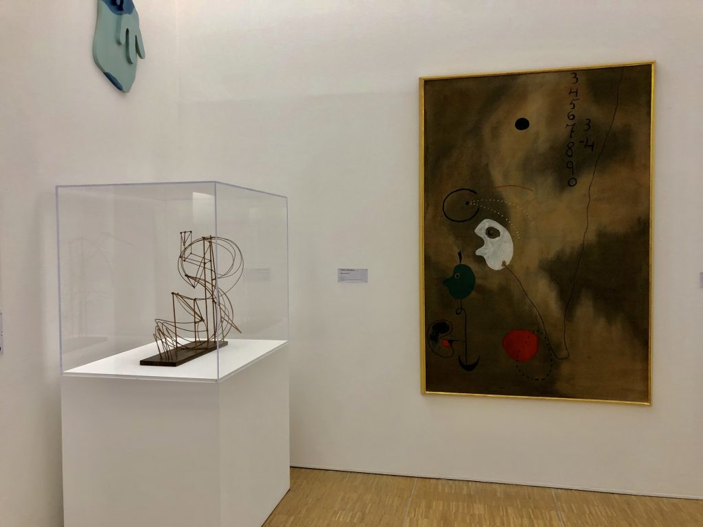A modern art museum display features a wire sculpture in a glass case on the left and an abstract painting with numbers and shapes on the right. The gallery walls are white with wooden flooring.