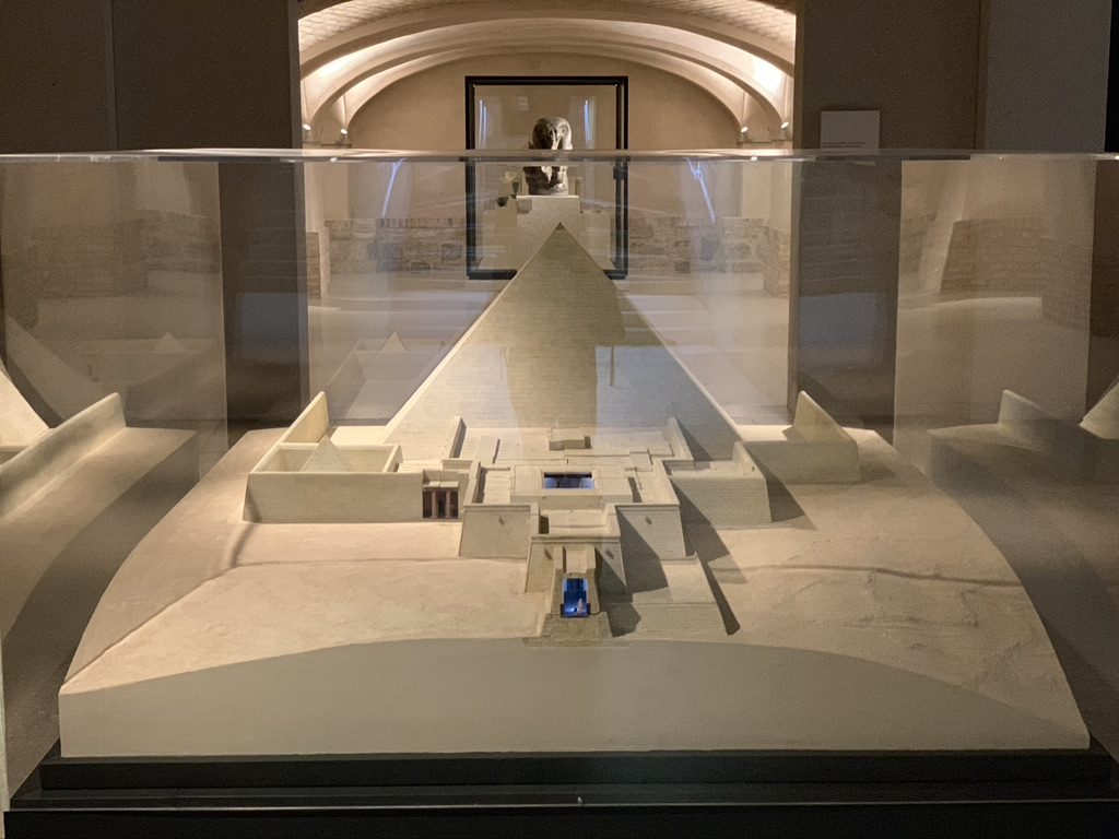 A scale model of an ancient pyramid, enclosed in glass. The model shows intricate architectural details with paths and surrounding structures. The display is set in a dimly lit room with arched ceilings and a framed image on the back wall.