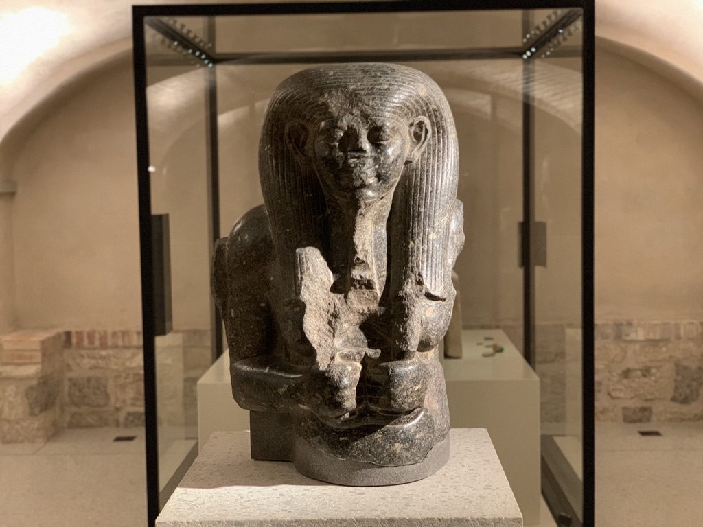 Ancient Egyptian statue displayed in a museum setting, enclosed in a glass case. The statue depicts a seated figure with a traditional headdress and a serene expression, holding objects. The background shows stone walls and dim lighting.