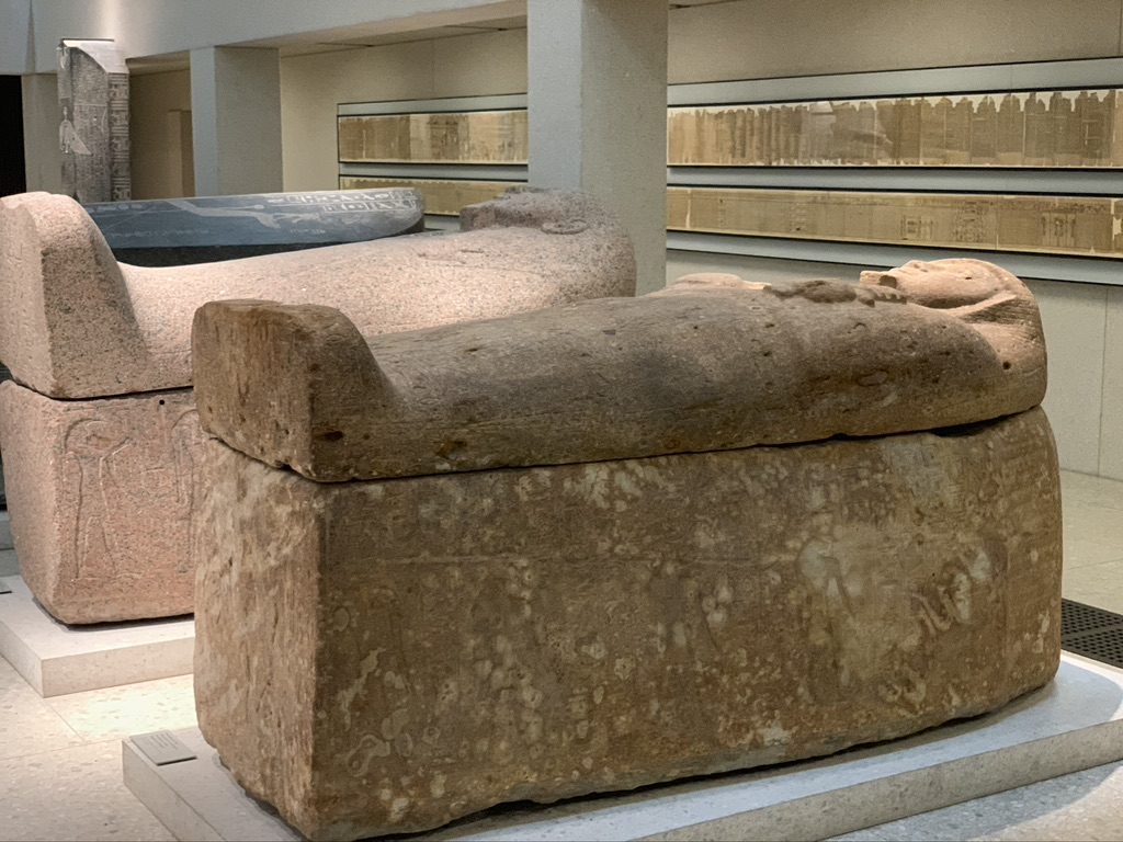 Two ancient stone sarcophagi displayed in a museum setting. The foreground shows a detailed, carved lid, while the background contains wall-mounted inscriptions and another sarcophagus. The scene is well-lit and captures the artifacts details.