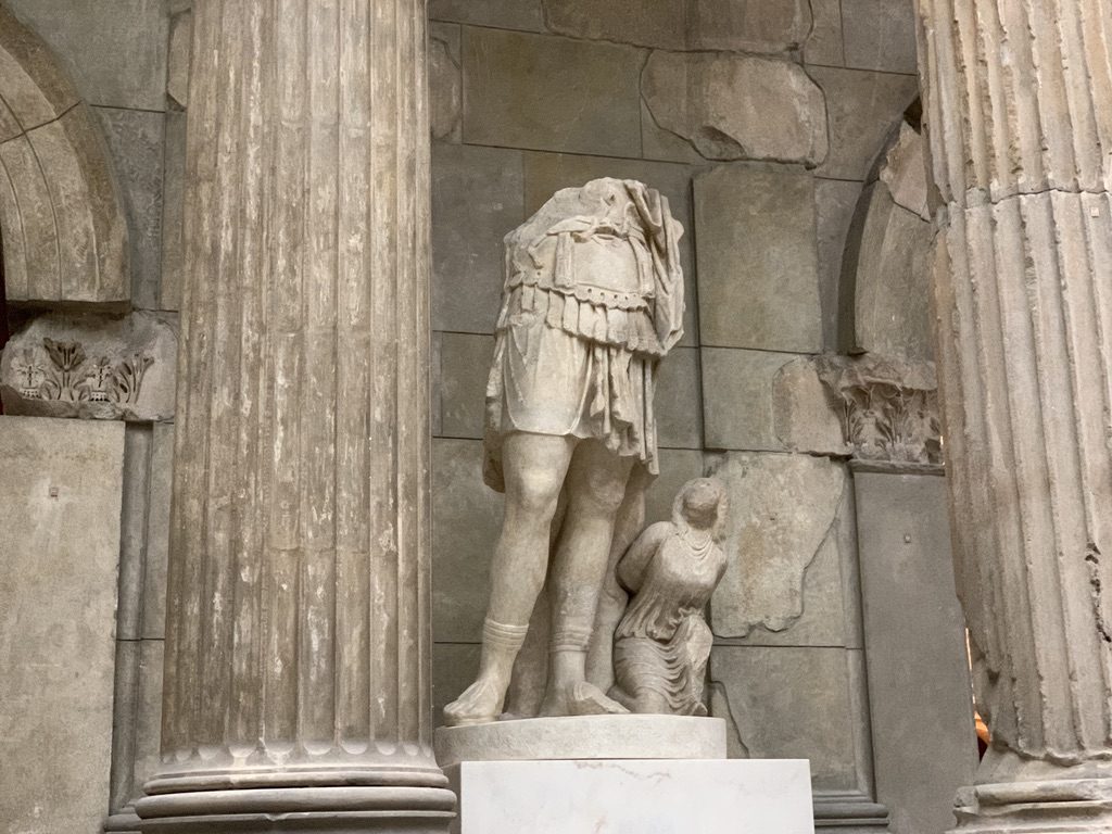 Ancient headless statue of a standing figure in a tunic, with a smaller kneeling figure beside it. The setting includes large stone columns and a textured stone wall.