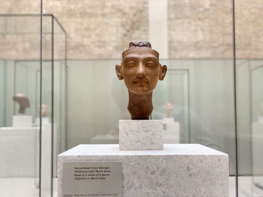 A stone bust of an Egyptian queen is displayed in a glass case within a museum setting. The sculptures neutral expression and detailed features stand out against a background of similar artifacts. The exhibit is well-lit, highlighting the artworks texture.