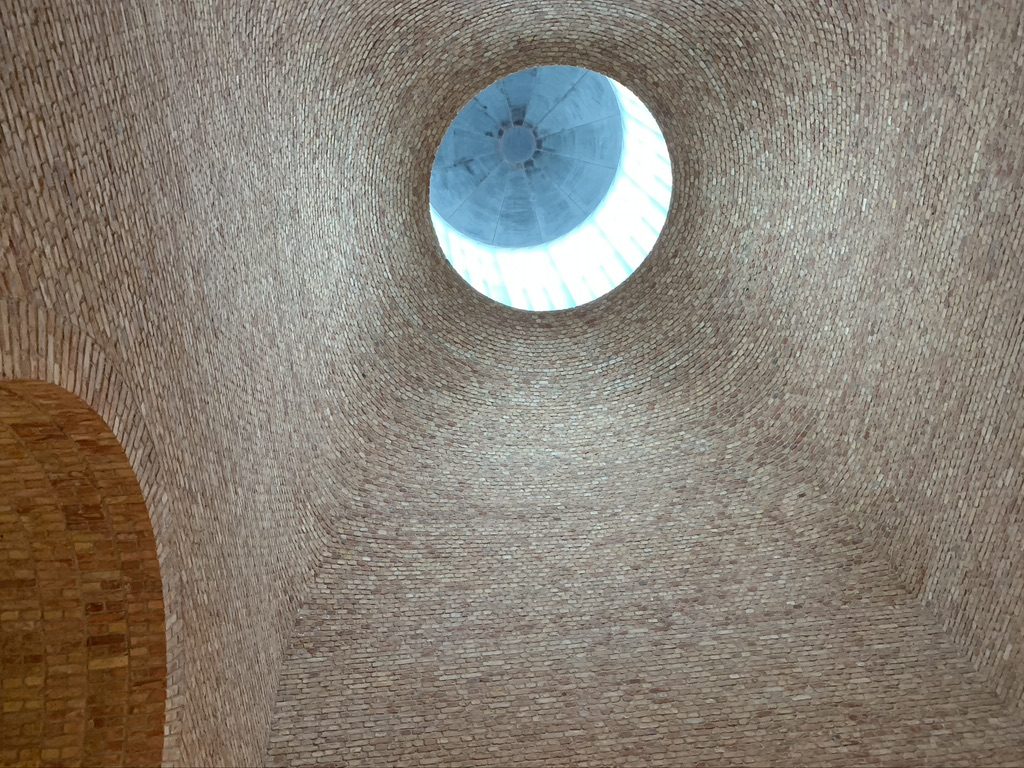 A brick dome interior with a circular skylight at the top, allowing natural light to enter. The textured brickwork creates a pattern leading up to the skylight, which provides a view of the blue sky above.