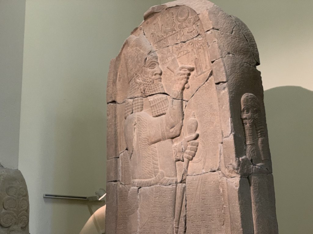 Ancient stone relief depicting two figures, one prominently holding a staff and wearing detailed attire. The carved figures are set against a smooth background, with intricate detailing on the clothing and symbols at the top.