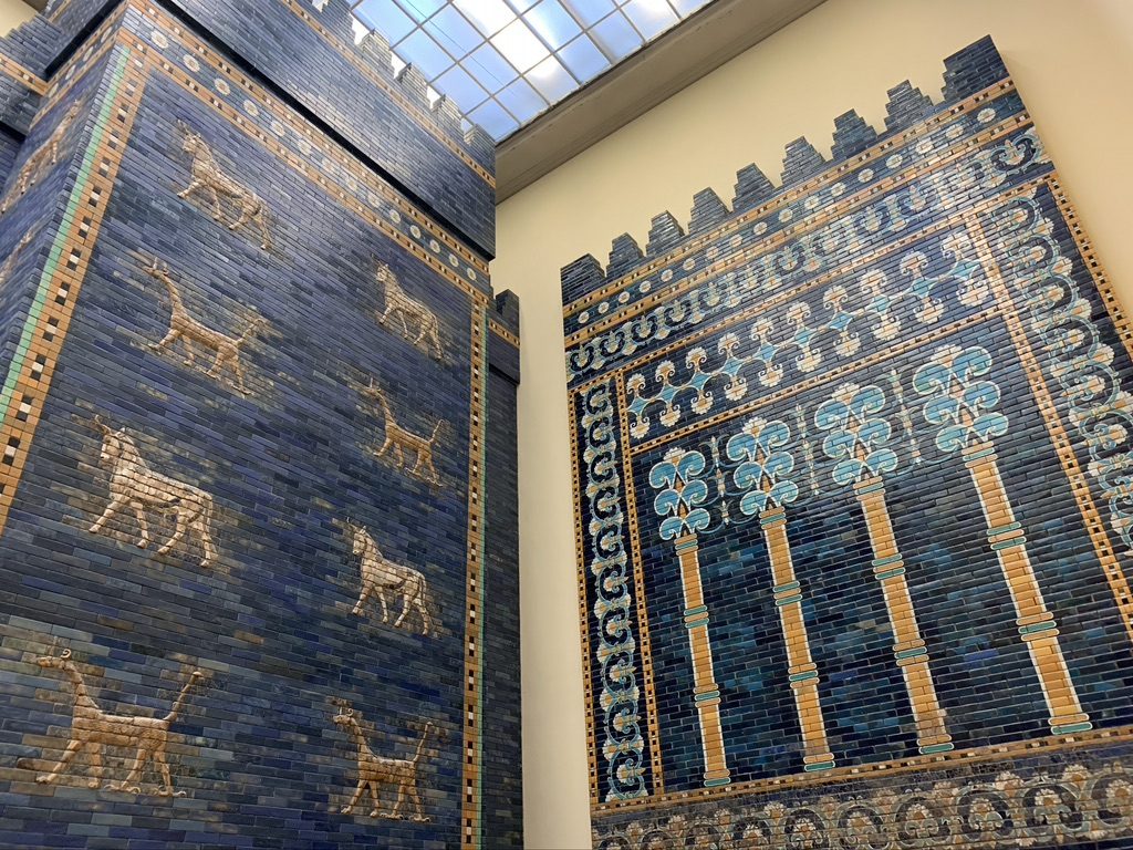 The image shows two large, ancient Babylonian walls covered in intricate blue and gold mosaics featuring mythical animal designs and decorative patterns. A glass roof provides overhead lighting.