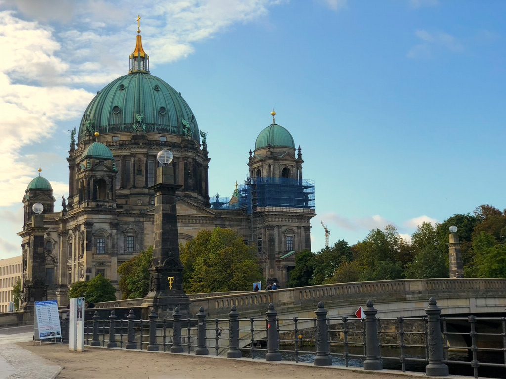 A historic cathedral with large domes stands near a bridge. The sky is partly cloudy. Green trees surround the building, and scaffolding is visible on one section of the structure. The foreground shows part of a stone balustrade along the river.
