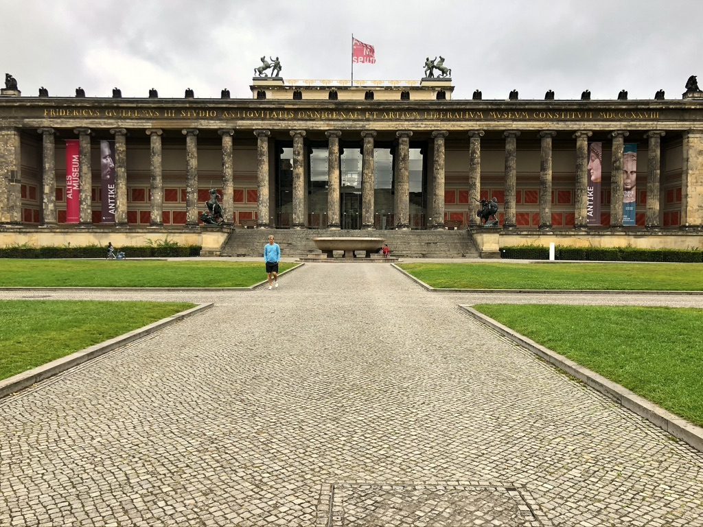 Exterior view of a neoclassical building with columns, banners, and a central entrance. A person walks on a path in the foreground, surrounded by green lawns. Overcast sky in the background.