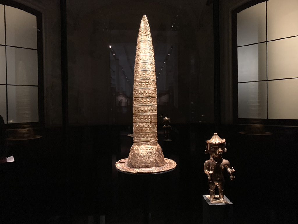 A tall, conical golden artifact with intricate patterns displayed in a dimly lit glass case. To its right, a small bronze figurine stands on a pedestal. Both objects are set against a backdrop of large, darkened windows.