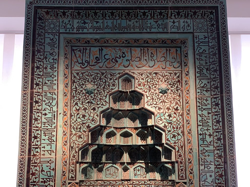 Intricately designed wall with ornate Arabic calligraphy surrounding a geometric, tiered niche pattern. The turquoise and earth-toned tiles create a detailed, symmetrical motif with floral and vine elements. Soft light highlights the texture and details.
