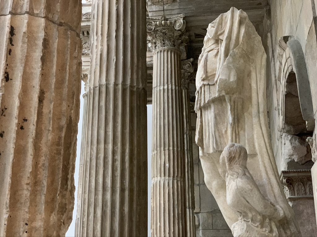 Ancient stone columns and statues, including a draped figure, inside a historic Greek or Roman structure. Weathered and detailed with carved patterns, reflecting classical architecture.