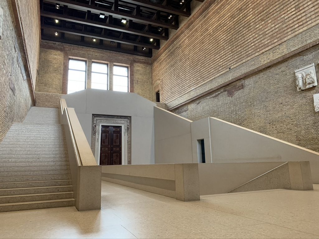 Interior view of a modern stairway and landing with minimalist design in a historic building. The area features exposed brick walls, large windows, and a wooden door. The ceiling has visible beams, creating an open and spacious atmosphere.