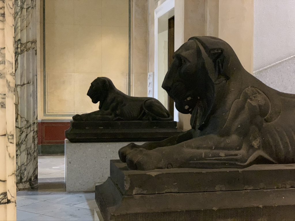 Two large stone lion sculptures are positioned on platforms in a hallway with marble columns. The lions are depicted in a reclining pose and face the same direction, adding a regal presence to the space.