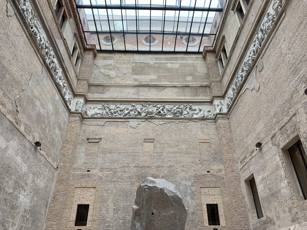 A large, rough-hewn stone stands in the center of a tall, rectangular room with brick walls. The room has a glass ceiling and features intricate sculptures lining the upper walls. Natural light streams in from above.