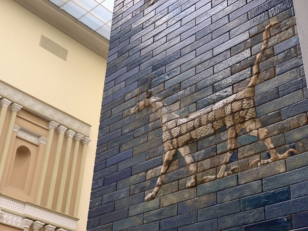A wall in a museum with blue bricks featuring a relief of a long-bodied animal. In the background, theres a beige structure with ornate decorative elements and a partially visible glass ceiling.