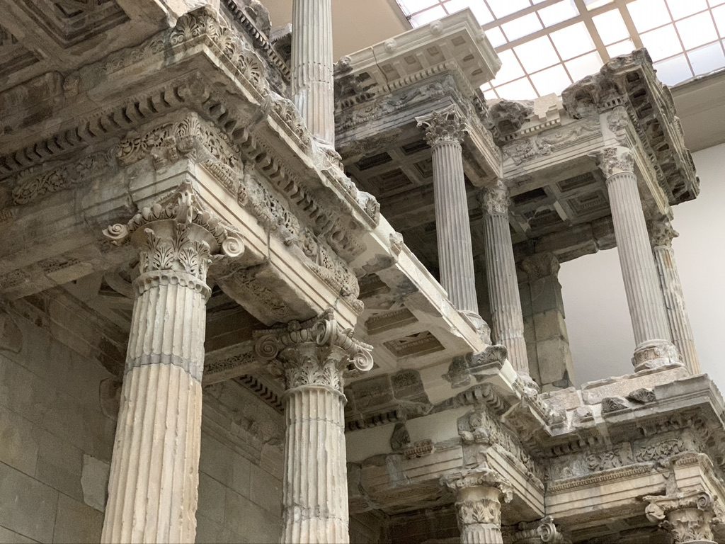 Ancient architectural ruins with detailed stone columns and intricate carvings against a skylight. The structure features multiple levels and ornate designs, showcasing historical craftsmanship.