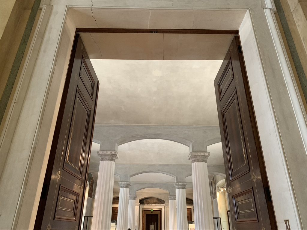 A grand entrance with wooden double doors leading to a building interior featuring tall, white columns and an ornate ceiling. The view captures an elegant, classical architectural design.