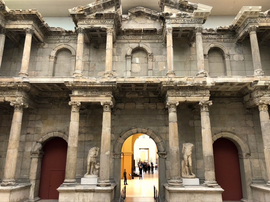 Ancient stone structure with columns and statues, featuring three arched doorways. Visitors are visible through one arch, walking inside a large hall with pale yellow walls, and a framed picture at the far end.