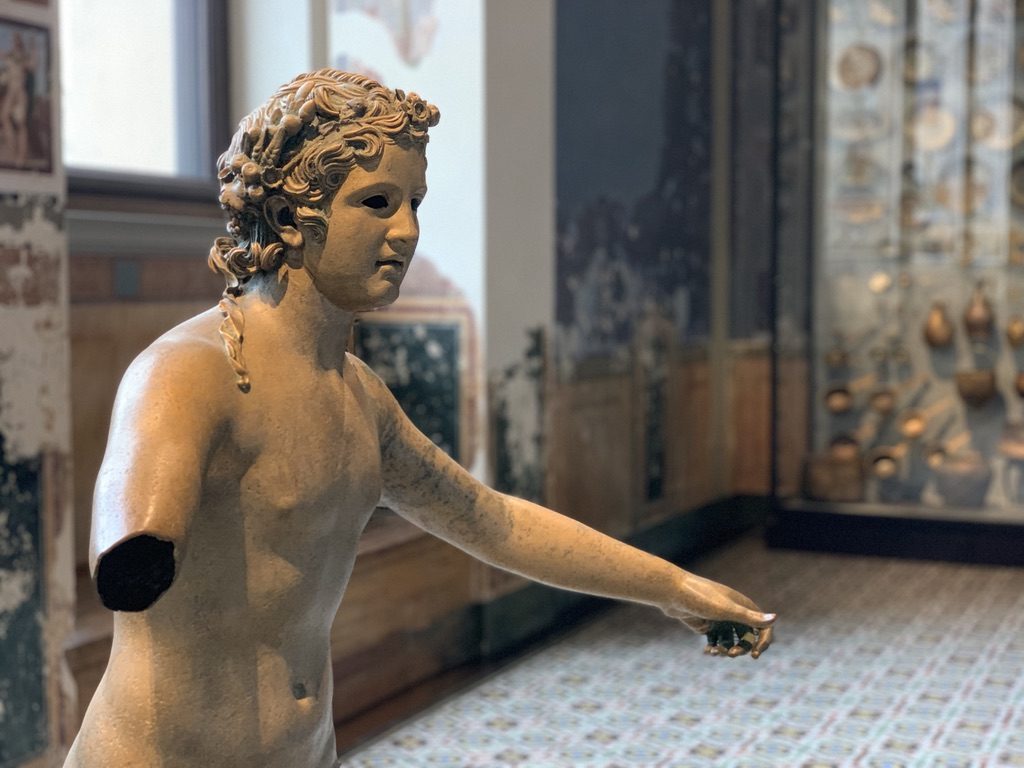 A close-up of a classical marble statue of a person with curls, missing both arms. The background shows a museum setting with artifacts displayed in a glass case and walls adorned with faded frescoes.