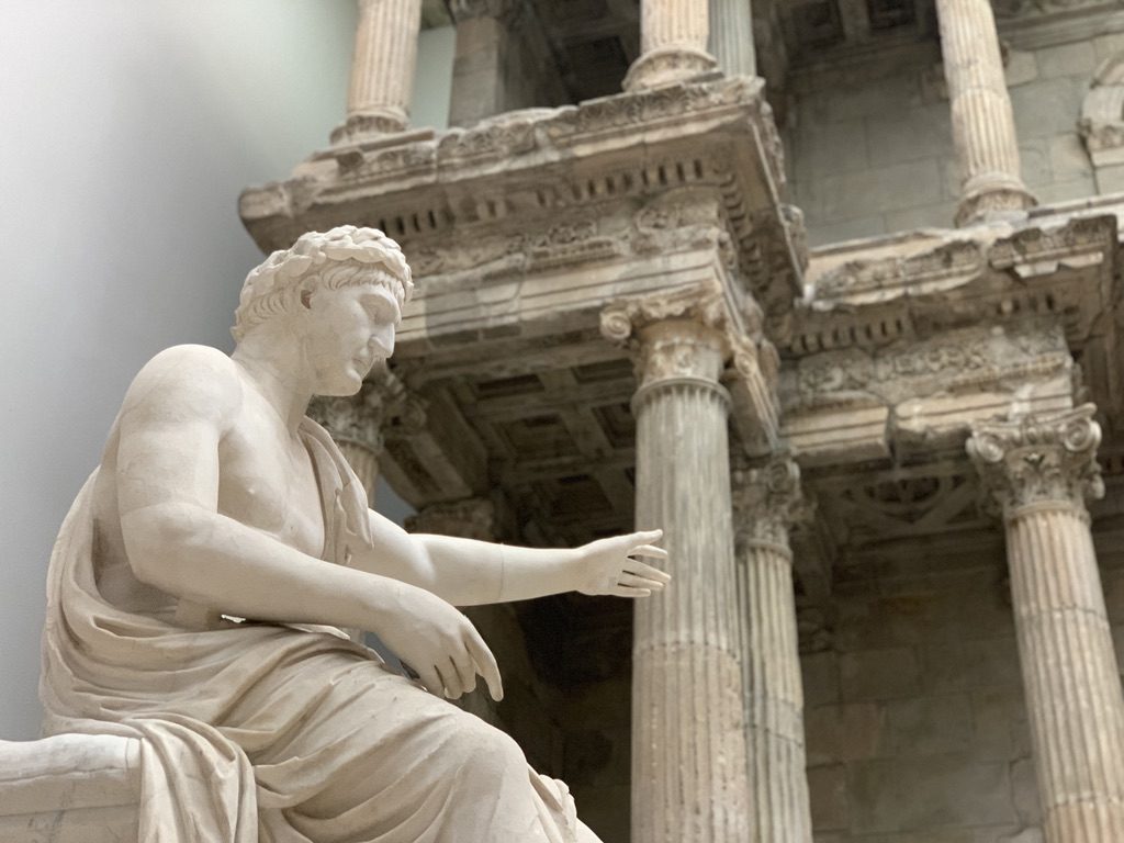 A marble statue of a seated figure with an extended arm in front of an ancient Roman-style architectural structure with tall, ornate columns and intricate details. The setting appears to be within a museum or historical site.
