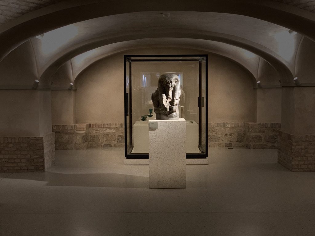 A dimly lit room with arched ceilings displays an ancient statue enclosed in a glass case. The statue, featuring a detailed anthropomorphic figure, stands on a pedestal in the center, surrounded by stone walls and sparse lighting.