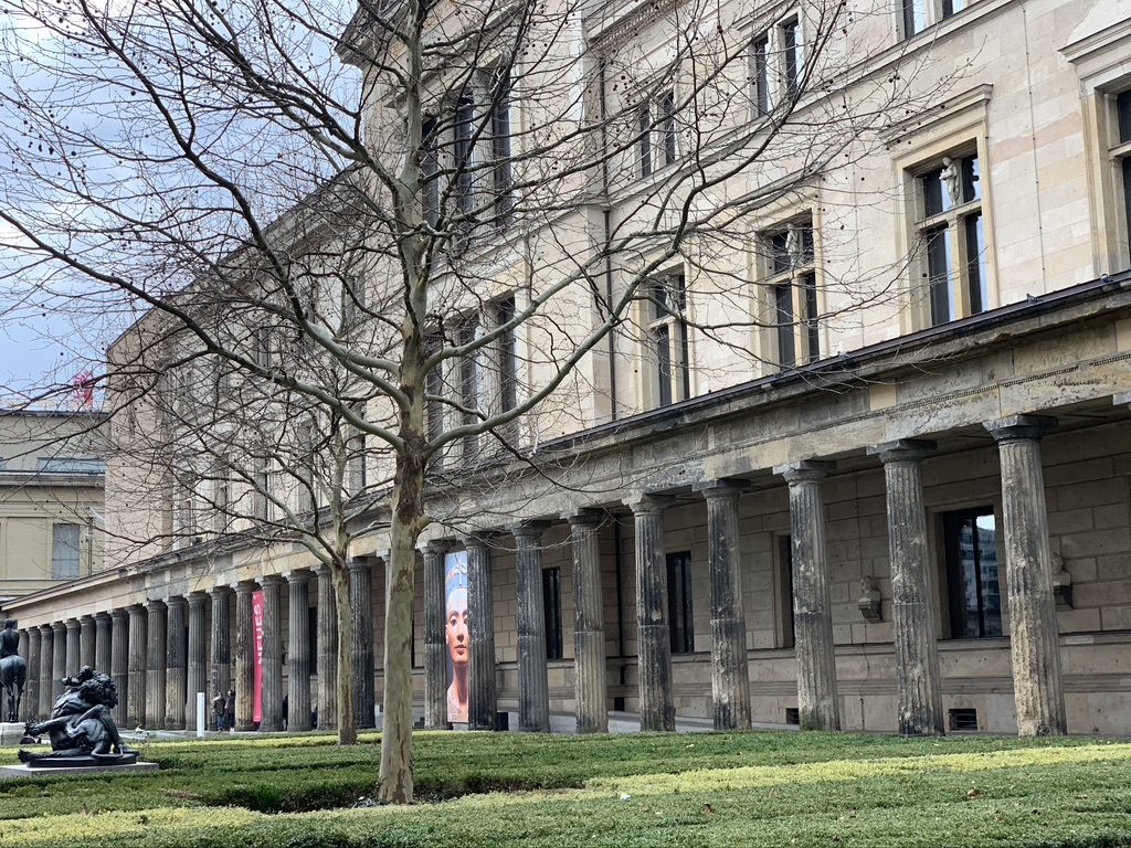 A historic building with tall columns and a barren tree in front. The architecture features multiple windows, and there is a colorful portrait banner hanging on one section. Green manicured hedges line the foreground.