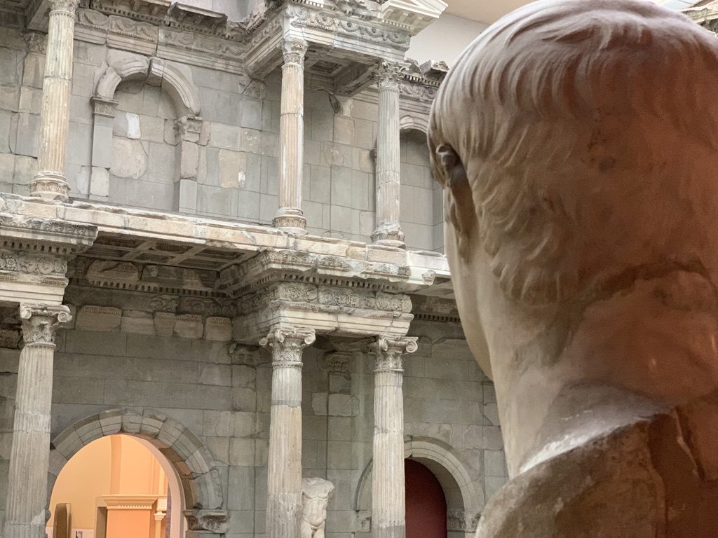 A marble bust viewed from behind, overlooking an ancient stone building with columns and arches inside a museum. The perspective highlights the intricate architectural details and historical ambiance.