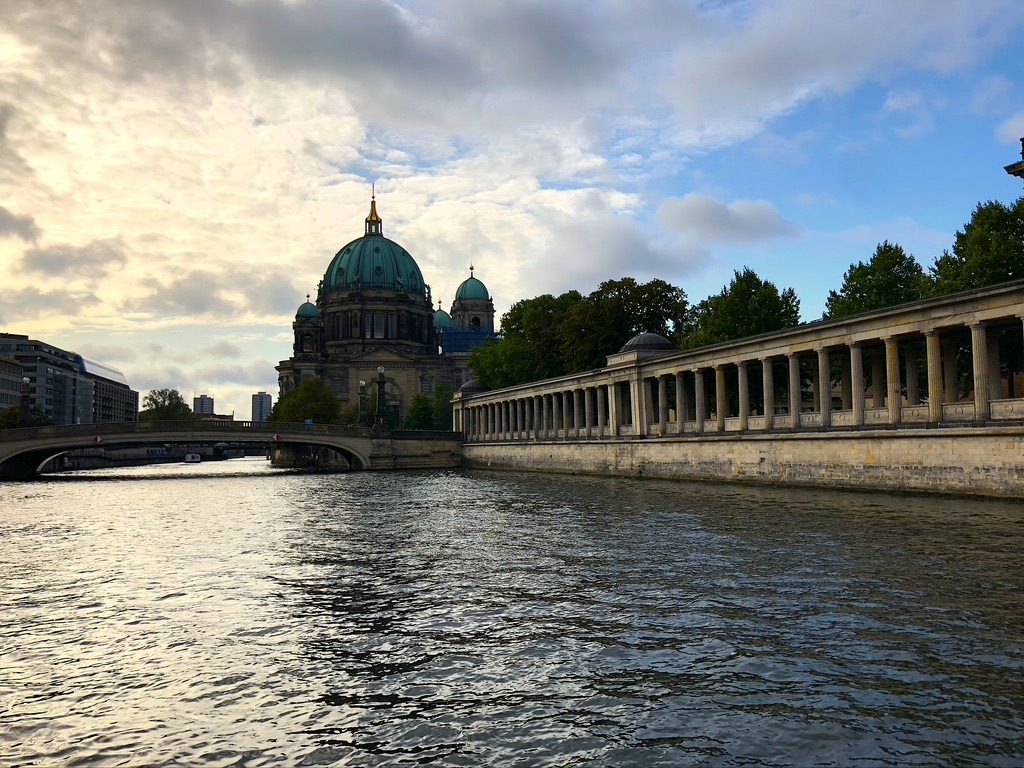 View of a river in a city with a historic domed building on the left, an elongated colonnade on the right, and a bridge crossing the water. Trees and clouds fill the background, with soft lighting from the setting sun.