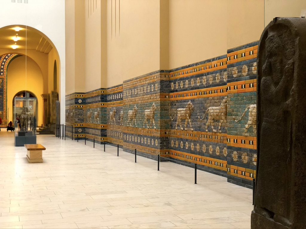 A long hallway in a museum showcases ancient Babylonian lion reliefs on a blue and brown tiled wall. The stone floor and arched ceilings create an expansive space. A bench and sculptures are visible in the distance.
