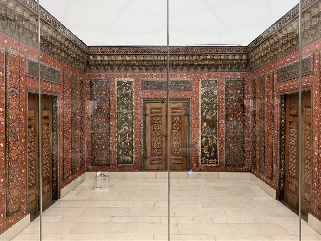 A lavishly decorated room with intricately painted walls, displaying detailed floral and geometric patterns in red, black, and gold. The room has wooden doors with ornate carvings and a cream-colored marble floor. A glass partition is visible.