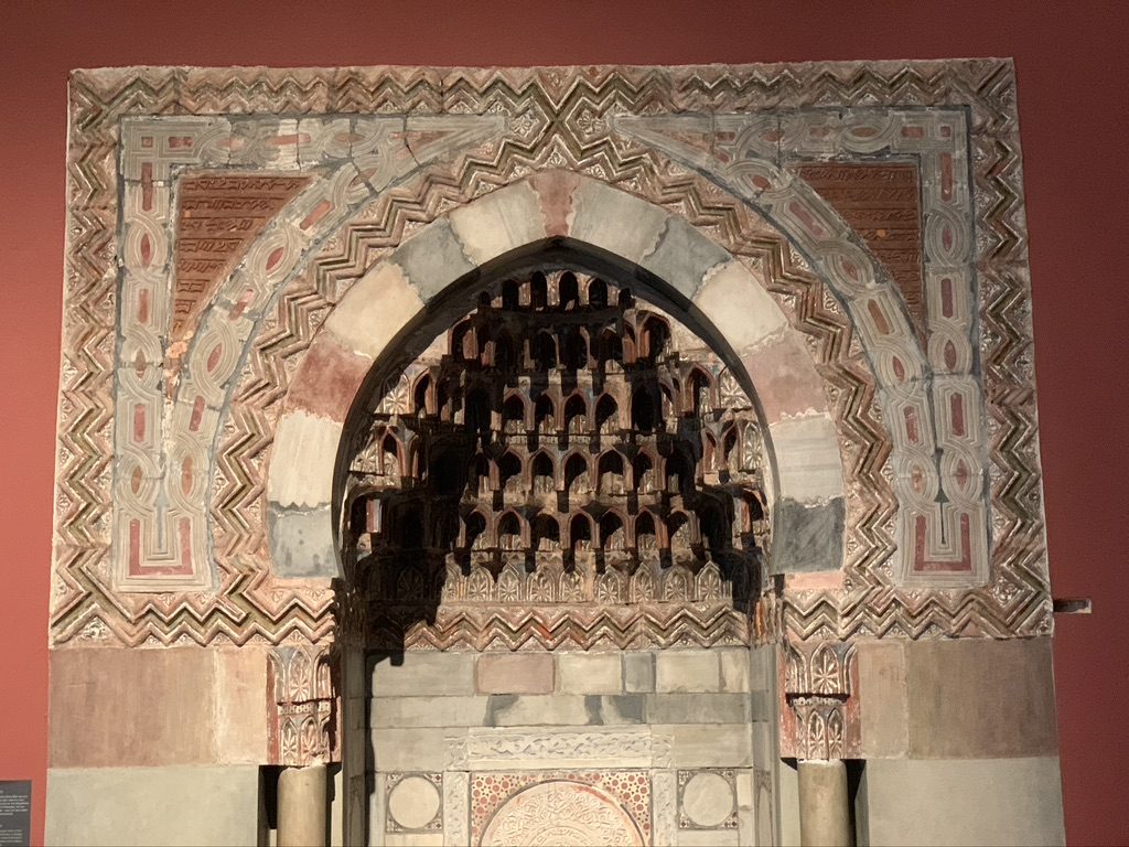 Intricate archway with decorative carvings and geometric patterns, featuring earthy tones of red, beige, and gray. The ornate designs highlight traditional architectural artistry against a rich red wall backdrop.