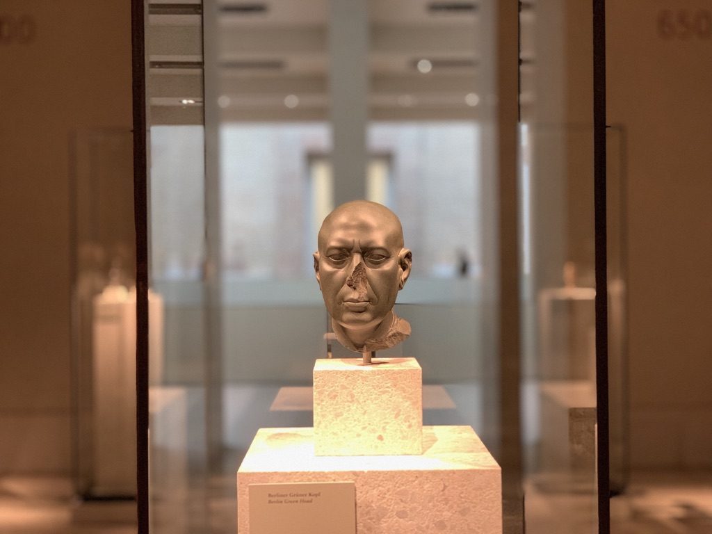 A sculpture of a human head is displayed on a pedestal in a museum setting. The exhibit is enclosed in a glass case, and the background is softly lit, creating a focused atmosphere on the artwork.