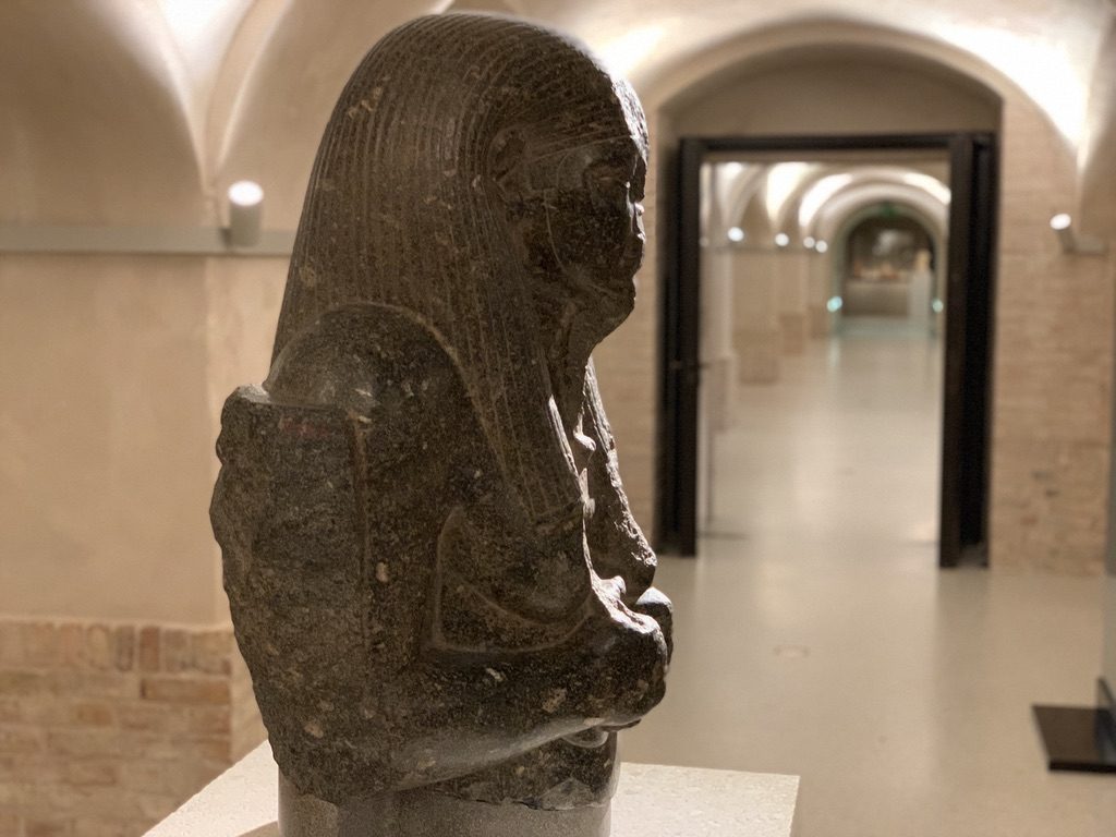 An ancient Egyptian stone statue of a seated figure with detailed carvings is displayed in a dimly lit museum hall with arched ceilings and brick walls in the background.
