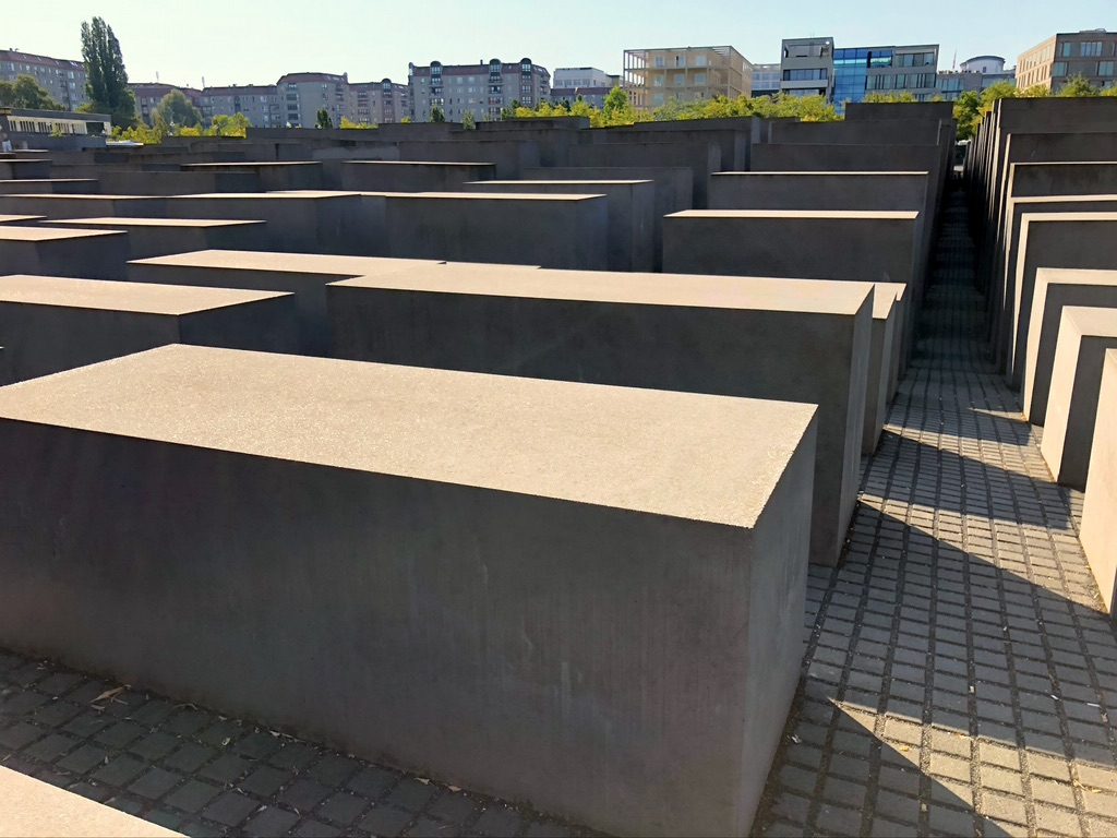 A large installation of concrete slabs of varying heights arranged in a grid pattern. The area appears to be an outdoor memorial or art installation, with a pathway between the slabs. Buildings and trees are visible in the background under a clear sky.