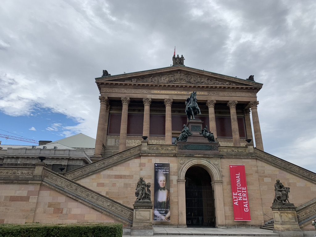 The image shows an imposing neoclassical building with grand staircases and a central archway. A statue of a rider on horseback is prominently displayed above the entrance. The sky is cloudy, and banners hang from the building facade.