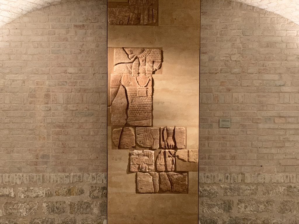 A wall-mounted, ancient stone relief depicting a standing figure with intricate details, surrounded by a stone brick archway. The relief is illuminated by soft lighting, highlighting the carving against the textured bricks.