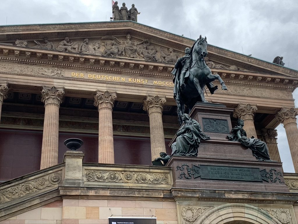 The image shows a historic building with classical columns and a detailed frieze. A bronze equestrian statue is prominently displayed in front, set against a cloudy sky. The building features the inscription Der Deutschen Kunst MDCCCLXXI.