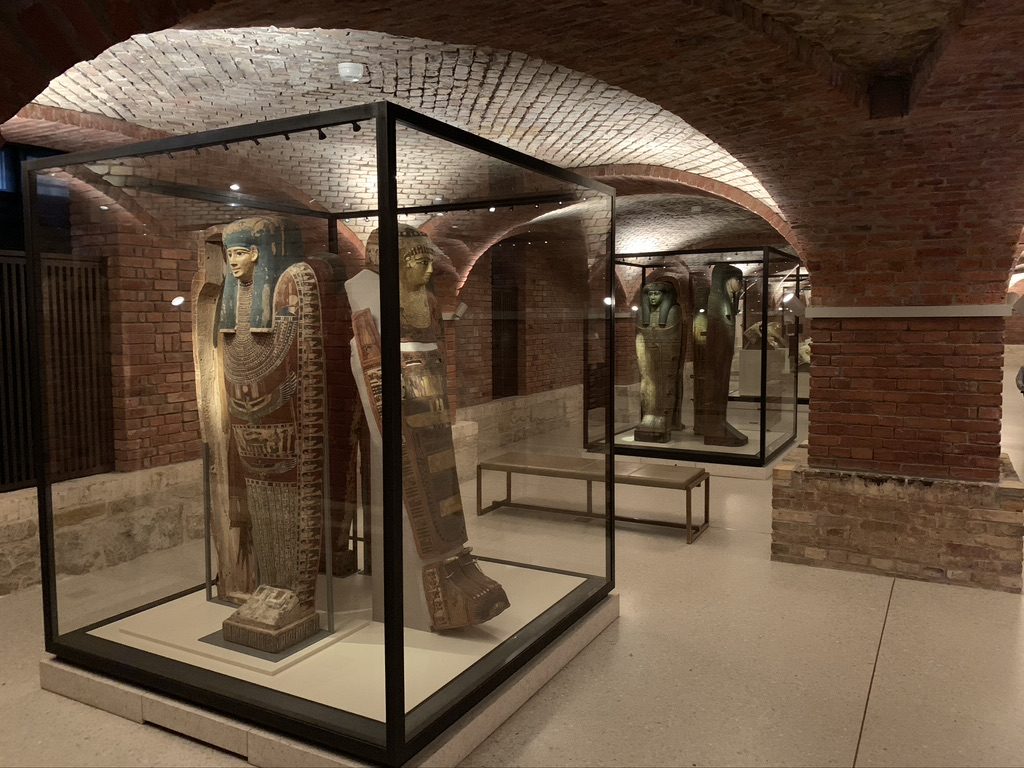 A museum gallery with Egyptian sarcophagi displayed in glass cases. The room has arched brick ceilings and warm lighting, creating a historic atmosphere. The sarcophagi are intricately detailed with ancient Egyptian motifs and symbols.