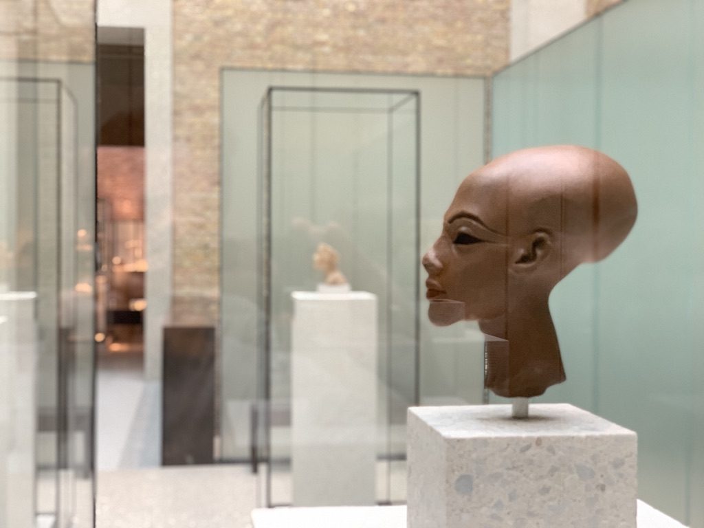 A museum display featuring an ancient Egyptian bust on a white pedestal. The bust, enclosed in a glass case, is set against a backdrop of a light-colored brick wall and other displayed artifacts in the background.
