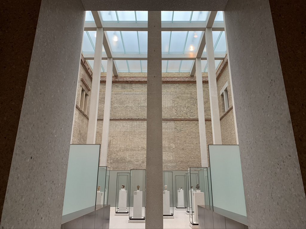 A modern exhibition space with glass displays containing artifacts. The room has high ceilings with skylights and is framed by a stone archway in the foreground. The walls are a mix of stone and brick.
