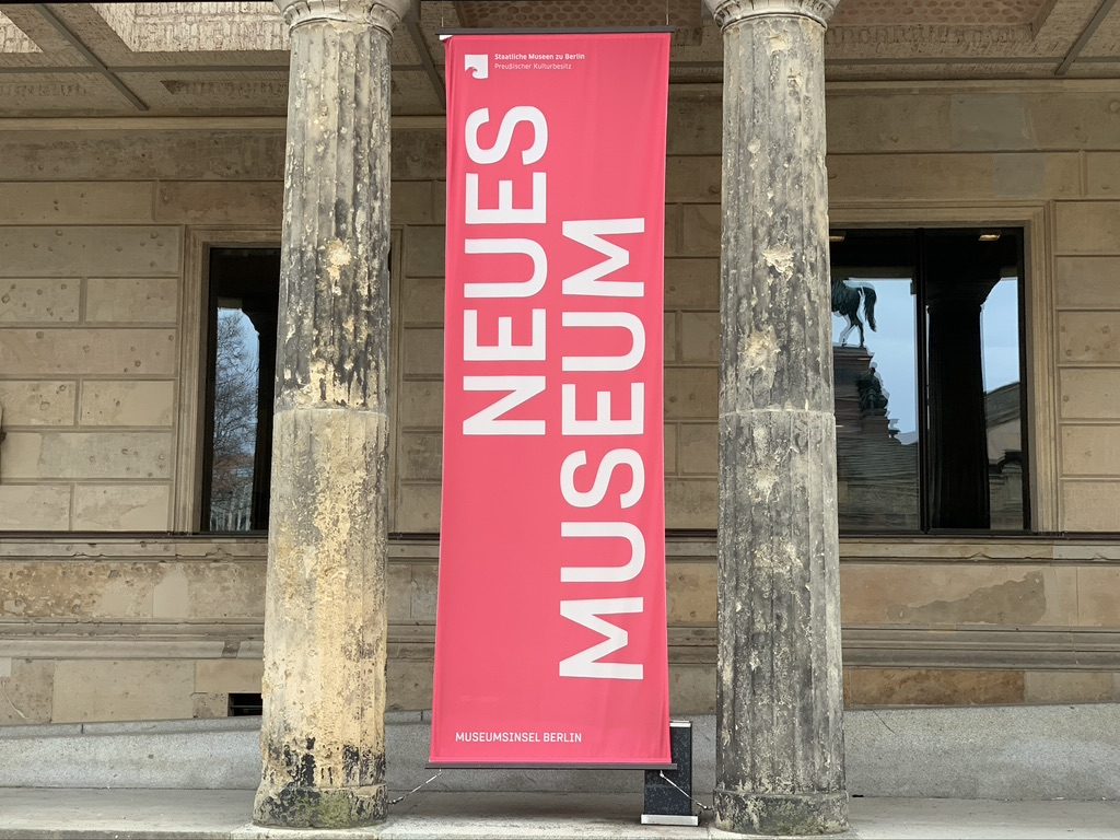 A tall pink banner with white text reads NEUES MUSEUM in front of a stone building with columns and windows. The setting is outdoors, and a statue is visible in the background through one of the windows.
