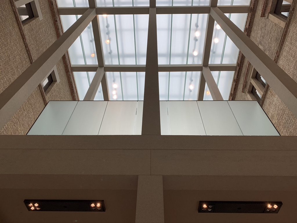 Upward view of an atrium ceiling with a grid-like pattern of large glass panels, allowing natural light. The walls are brick, and there are minimalist design elements, including frosted glass panels on the upper level.