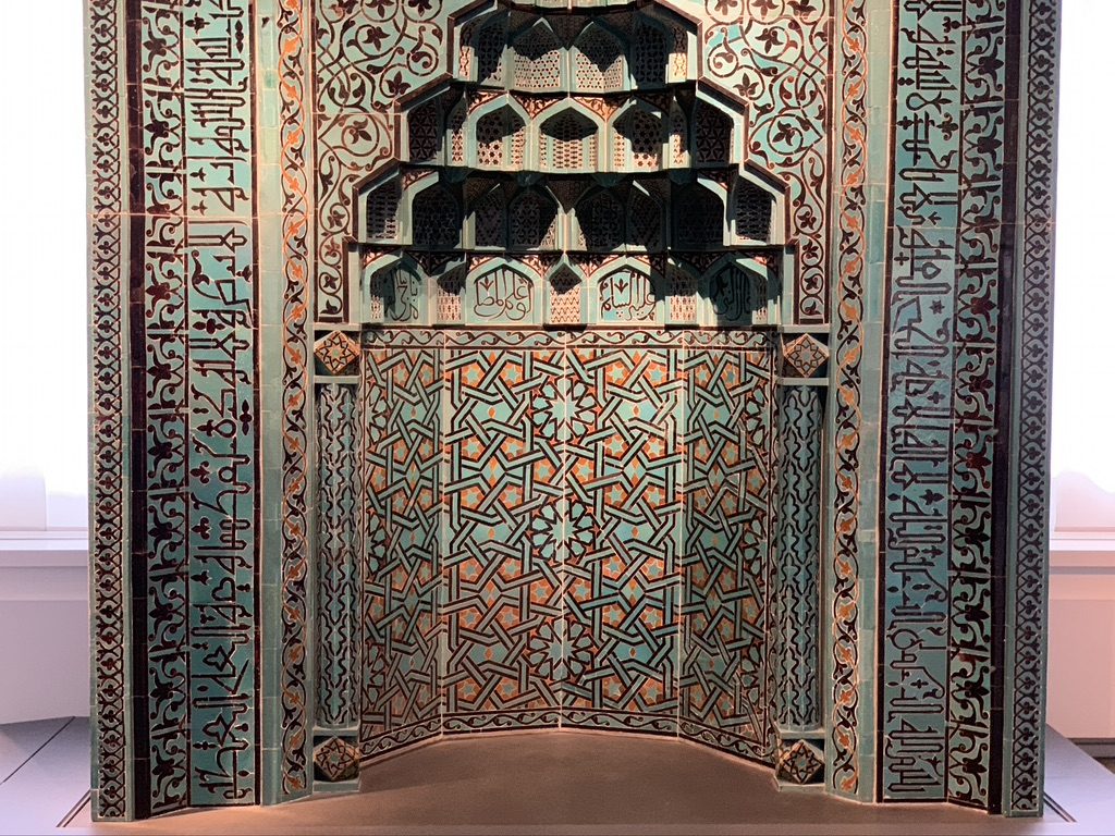 Intricate tile mosaic featuring geometric patterns and Arabic script in blue and brown hues, adorning a decorative wall panel.