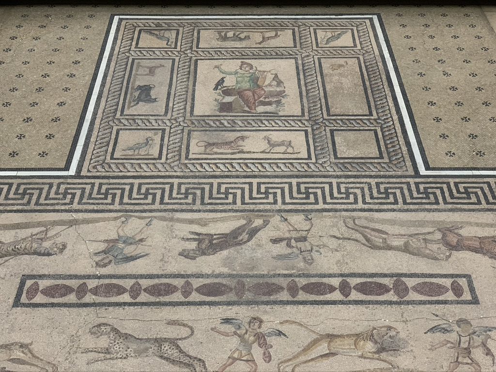 Ancient Roman mosaic floor with detailed patterns, showcasing central figures and various animals, including a depiction of lions. The intricate design includes geometric motifs and a border of interlocking lines.