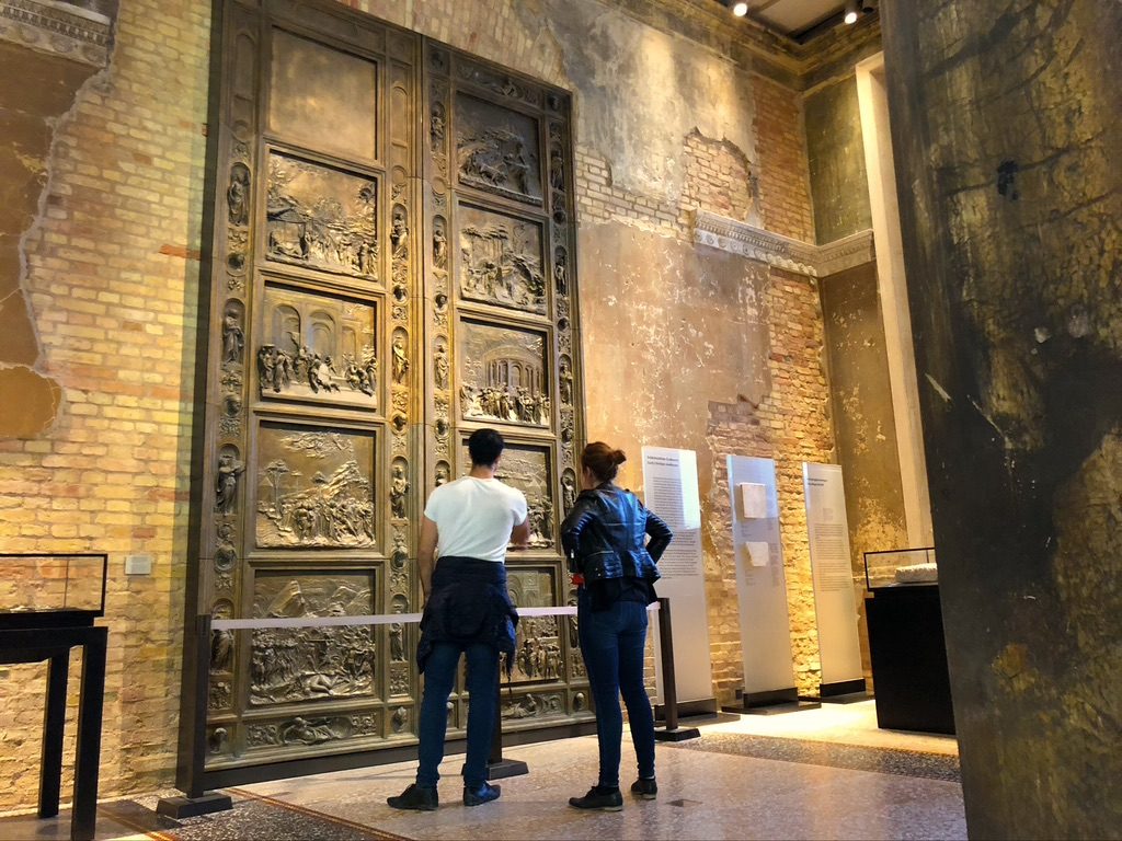 Two people stand in an art gallery, observing a large, ornate bronze door with intricate relief sculptures. Walls are exposed brick, and the setting is well-lit, highlighting the artwork’s details.